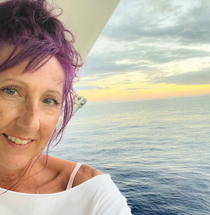 Portrait of Keri Dangerfield Stone with a sunset over the ocean in the background. She has vibrant purple hair and is smiling at the camera.